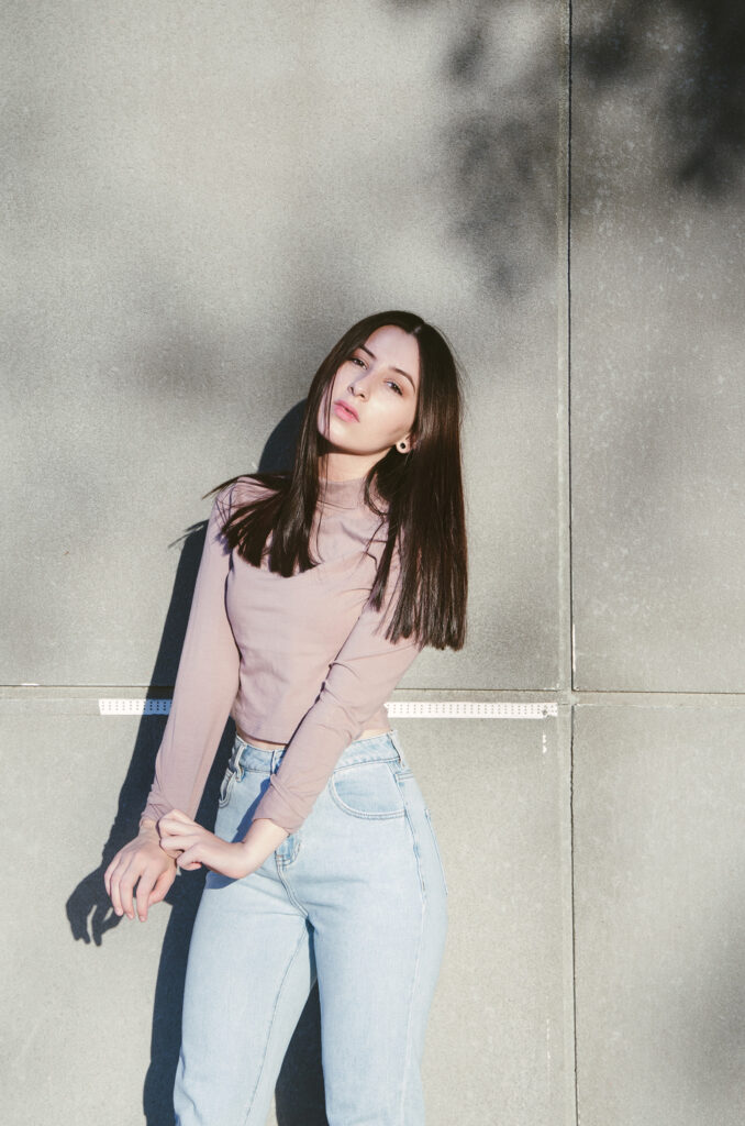Gabriela Inzunza poses against a gray wall casting a shadow wearing a mauve top and light blue jeans.